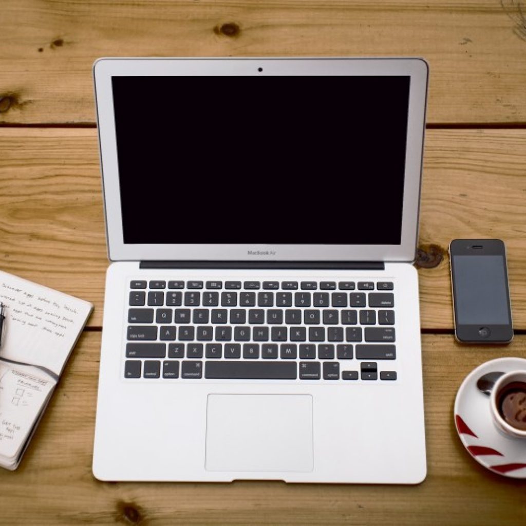 A laptop, notebook, mobile and teacup laying on top of a wooden slatted desk