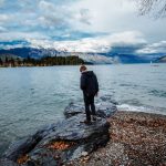 A person standing on a rock on the edge of a lake with a foest and mountain view in the background