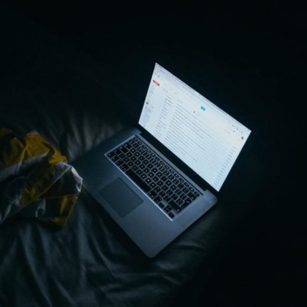 A laptop displaying an email inbox, lying on a bed in the dark