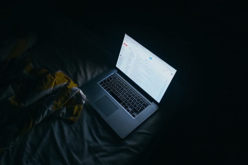 A laptop displaying an email inbox, lying on a bed in the dark