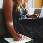 A woman typing on a laptop placed on her knee while writting in a notepad