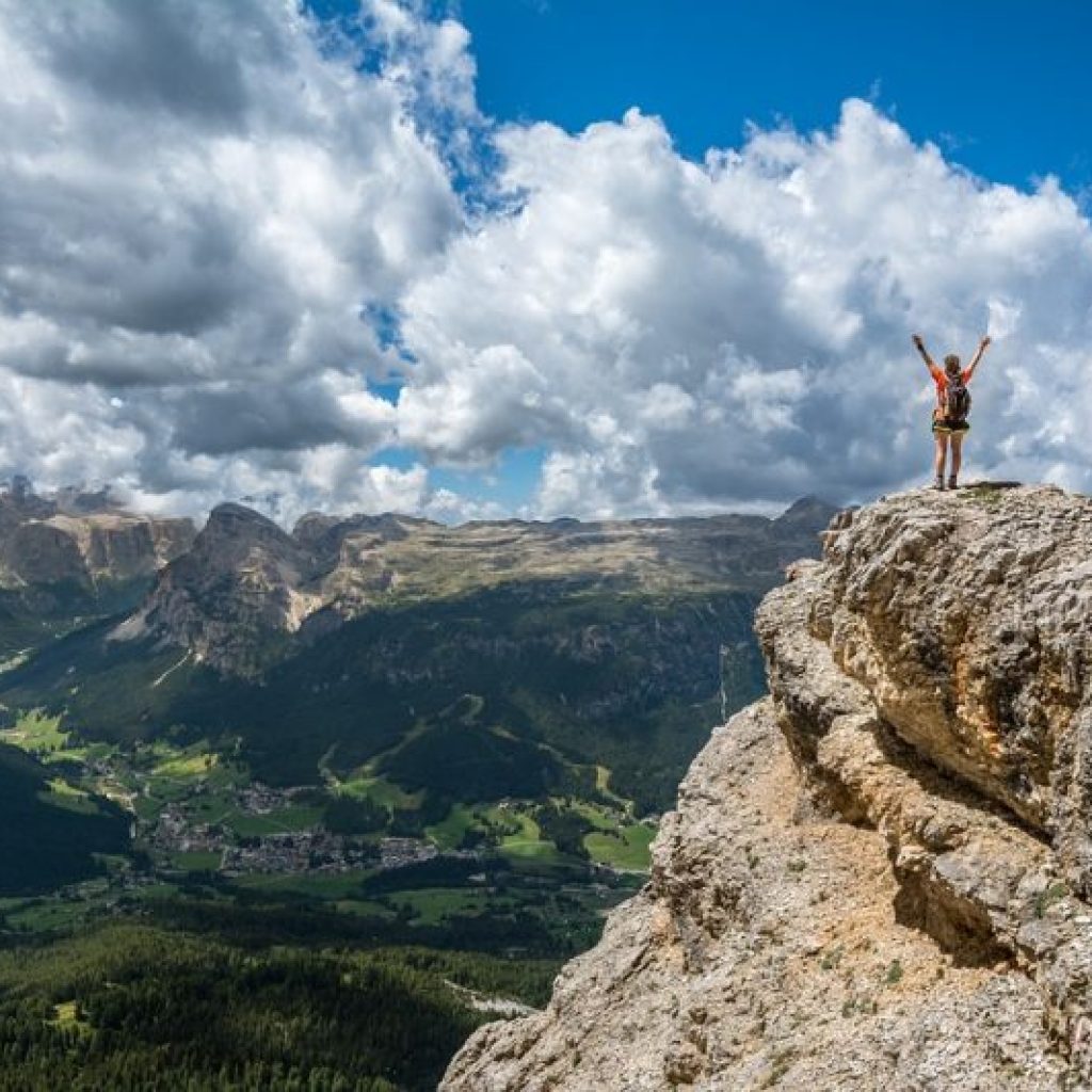 A mountain climber standing on top of a mountain peak celebrating with their hands up while overlooking a valley view