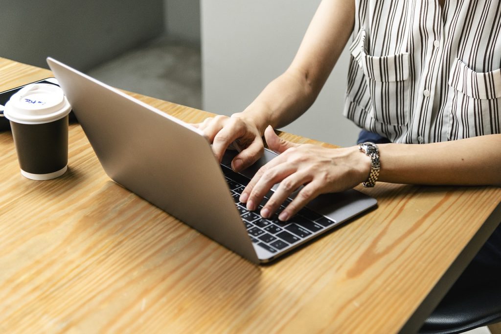 Woman in a white stripy top typing on a laptop while sat at a wooden desk with a coffe cup