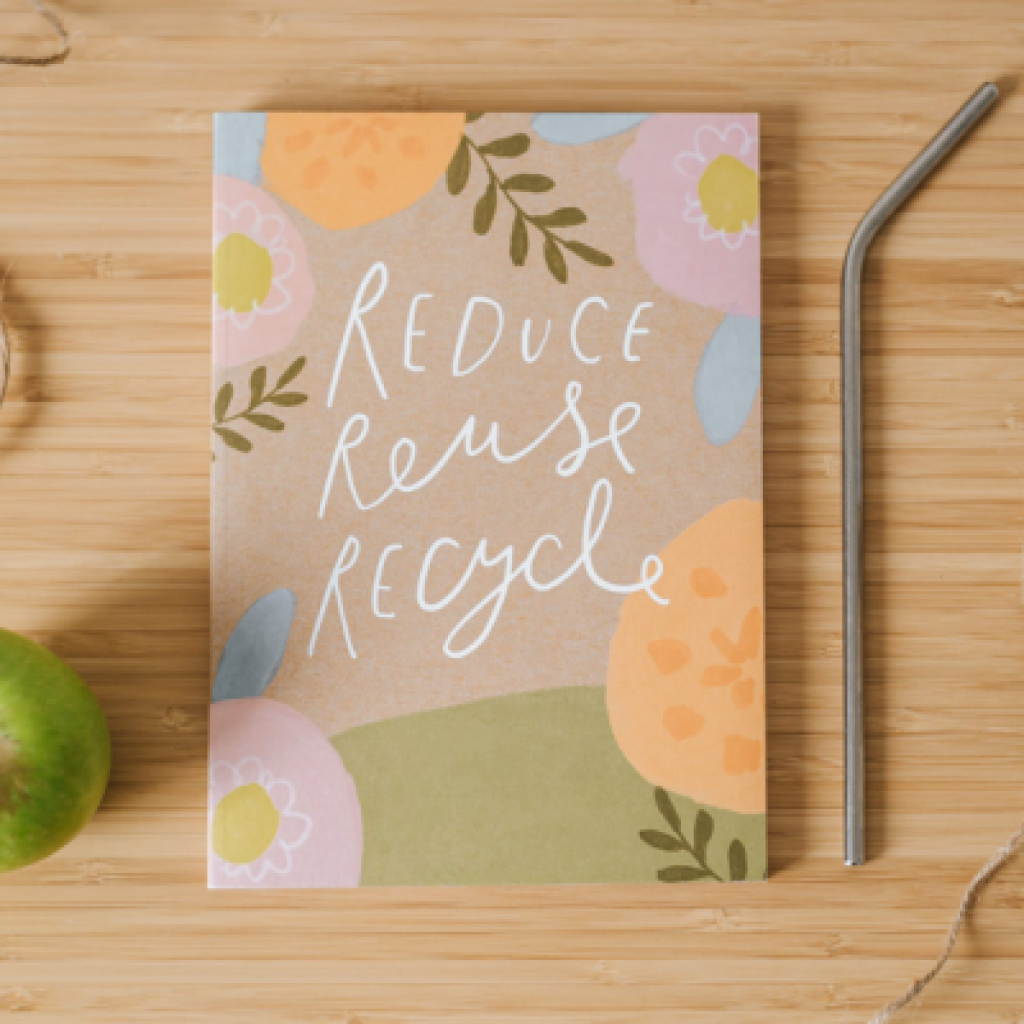 A notebook saying Reduce Reuse Recycle on the cover on top of a wooden surface alongside a green apple, some twine and a metal straw