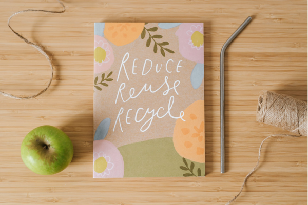 A notebook saying Reduce Reuse Recycle on the cover on top of a wooden surface alongside a green apple, some twine and a metal straw