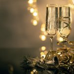 Two champagne glasses tanding on golden decorations and Christmas bells with warm lights in the background