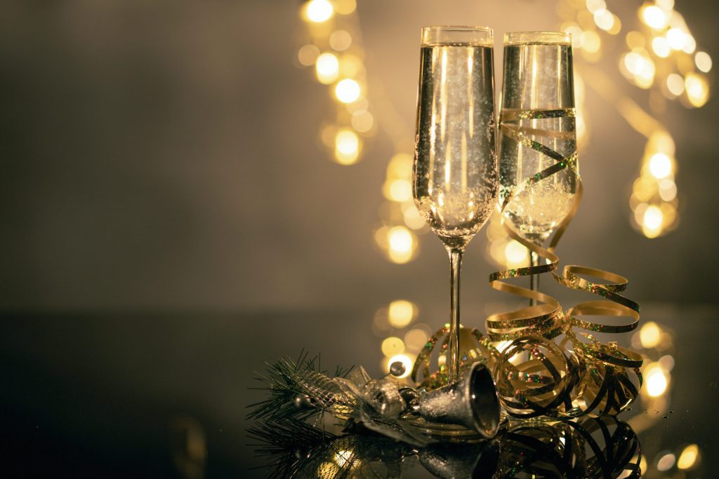 Two champagne glasses tanding on golden decorations and Christmas bells with warm lights in the background