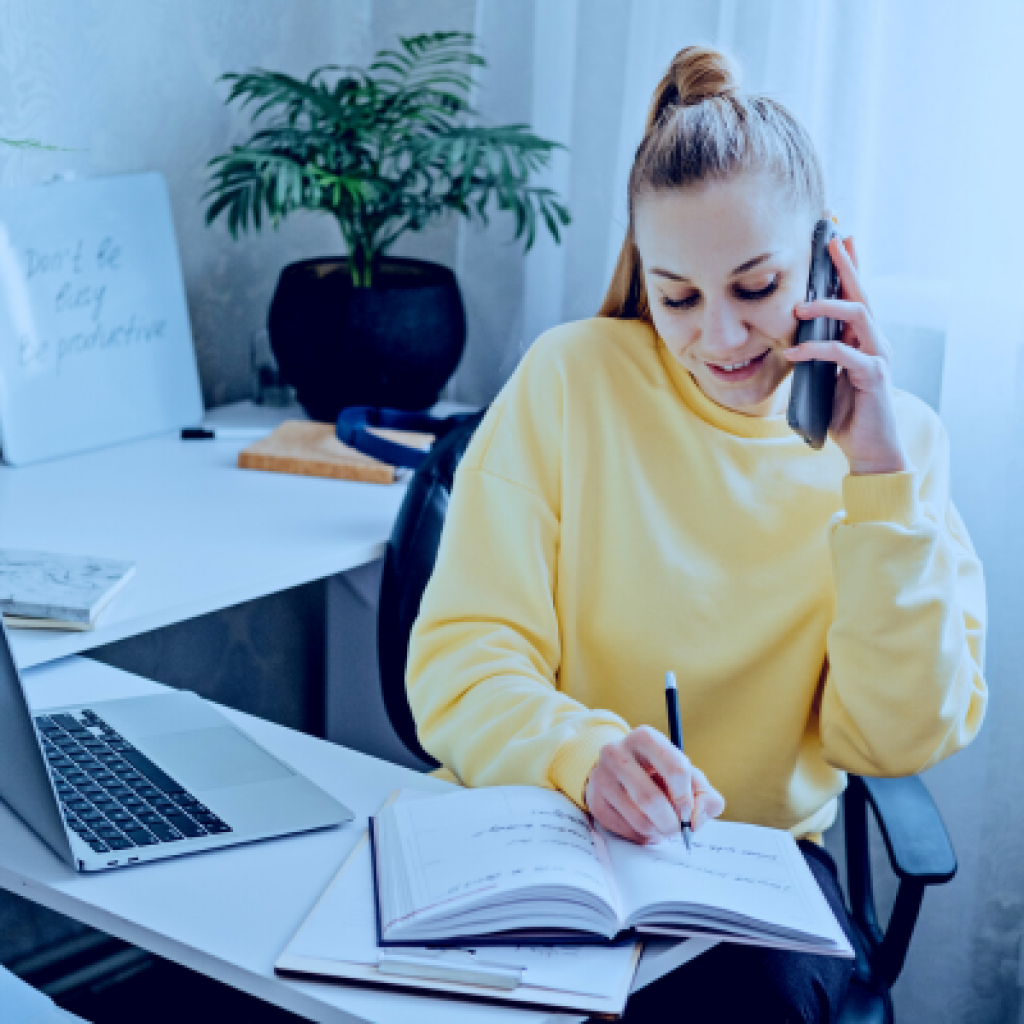 A woman working at a computer while taking a phonecall and writing in a notebook