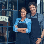 Two people standing infront of their small business while leaning against a door with a sign which reads "Open"
