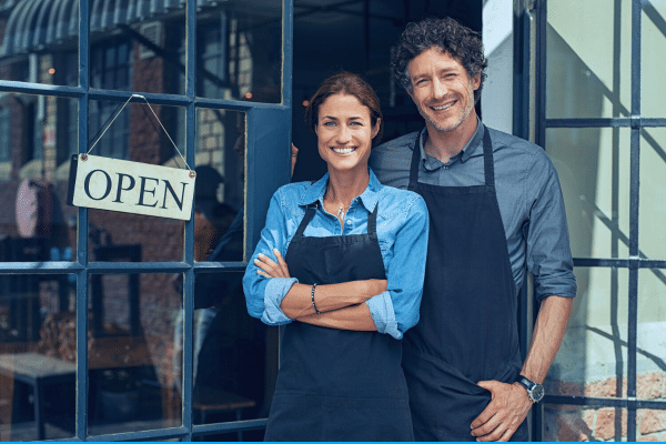 Two people standing infront of their small business while leaning against a door with a sign which reads "Open"