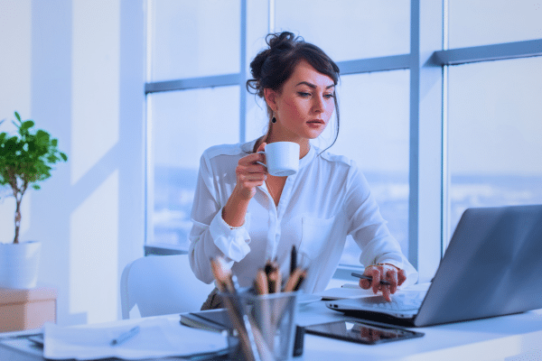 A woman in formal clothing working at a desk with an open laptop and a cup in her hand