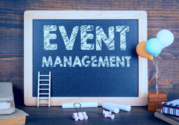 A chalkboard sign displaying the words "Event Management" in white writting with some decorative balloons and a small ladder